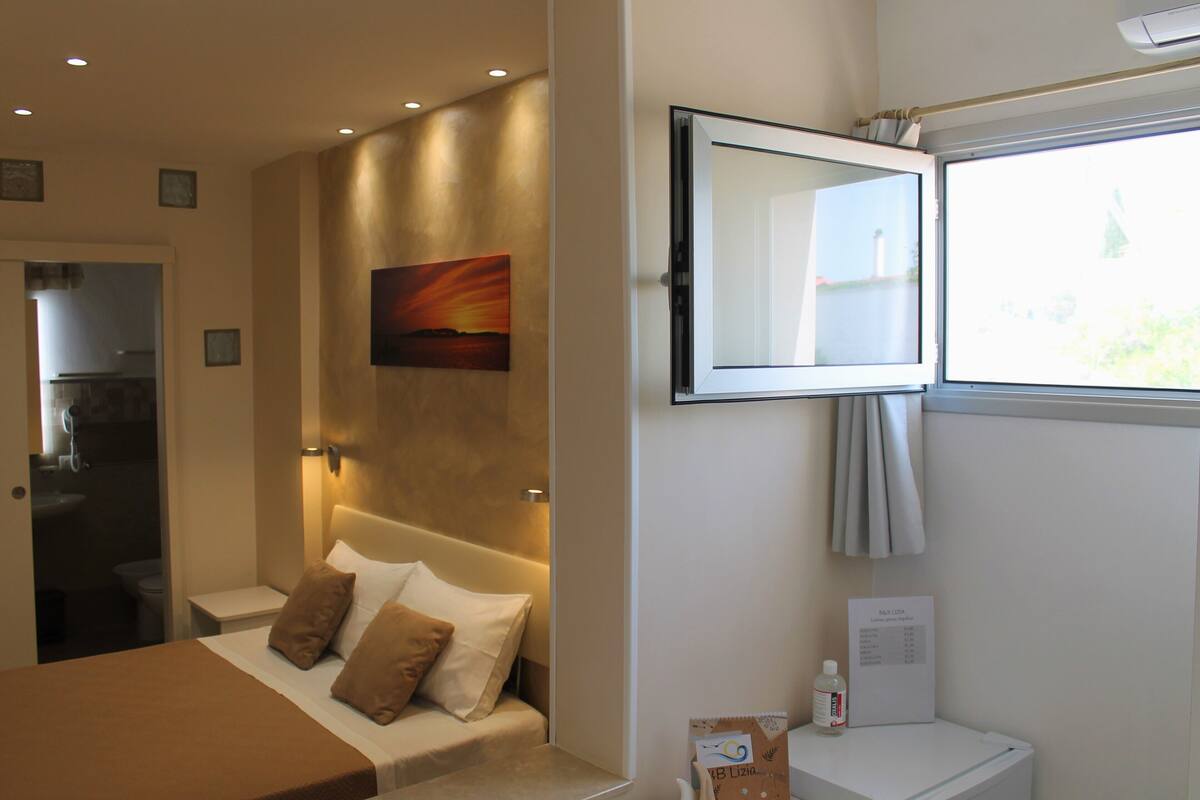 A hotel room with a bed, a TV mounted on the wall, a window with a view, and a bathroom visible through an open door.