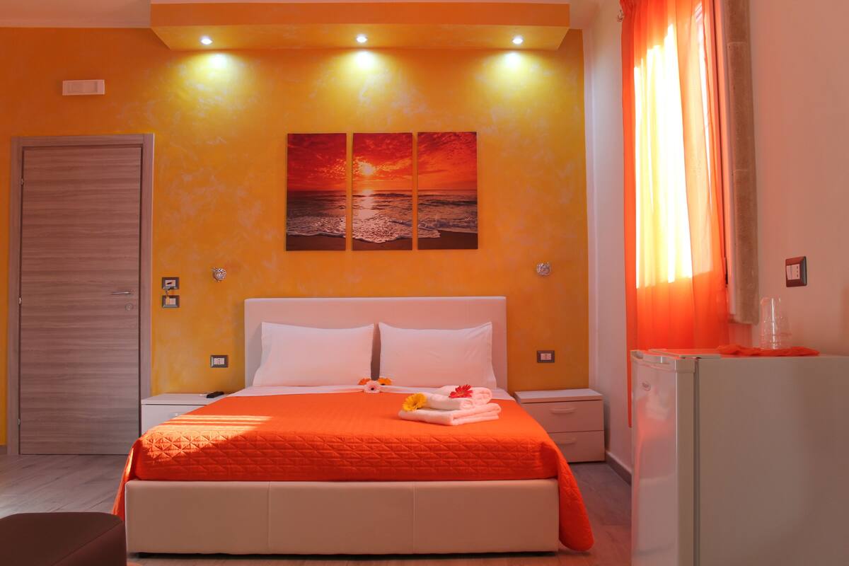A hotel room with a large bed, a nightstand, a dresser, and a painting of a sunset over the ocean.