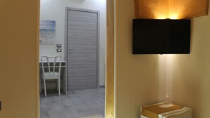 Ein Hotelzimmer mit geschlossener Tür, einem weißen Stuhl, einem an der Wand befestigten Flachbildfernseher und einem kleinen Tisch mit einer Flasche und einem Notizblock.