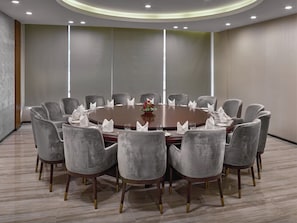 Un salón de conferencias con una mesa redonda, sillas tapizadas de gris y un florero con flores rojas.