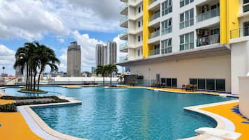 Una piscina con un paseo circundante, palmeras y un edificio moderno con balcones al fondo.