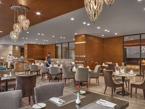 A modern dining area with multiple tables and chairs, a chef preparing food, and a large chandelier. - TUI SUNEO Olympia Phnom Penh (Phnom Penh)