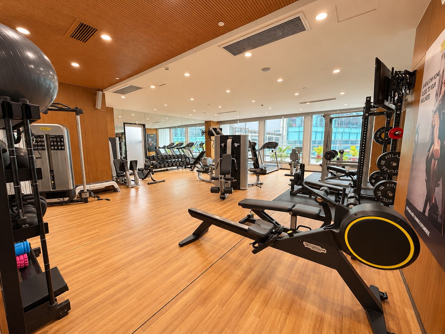 Un gimnasio con diferentes equipos de ejercicio, ventanas amplias y suelo de madera.