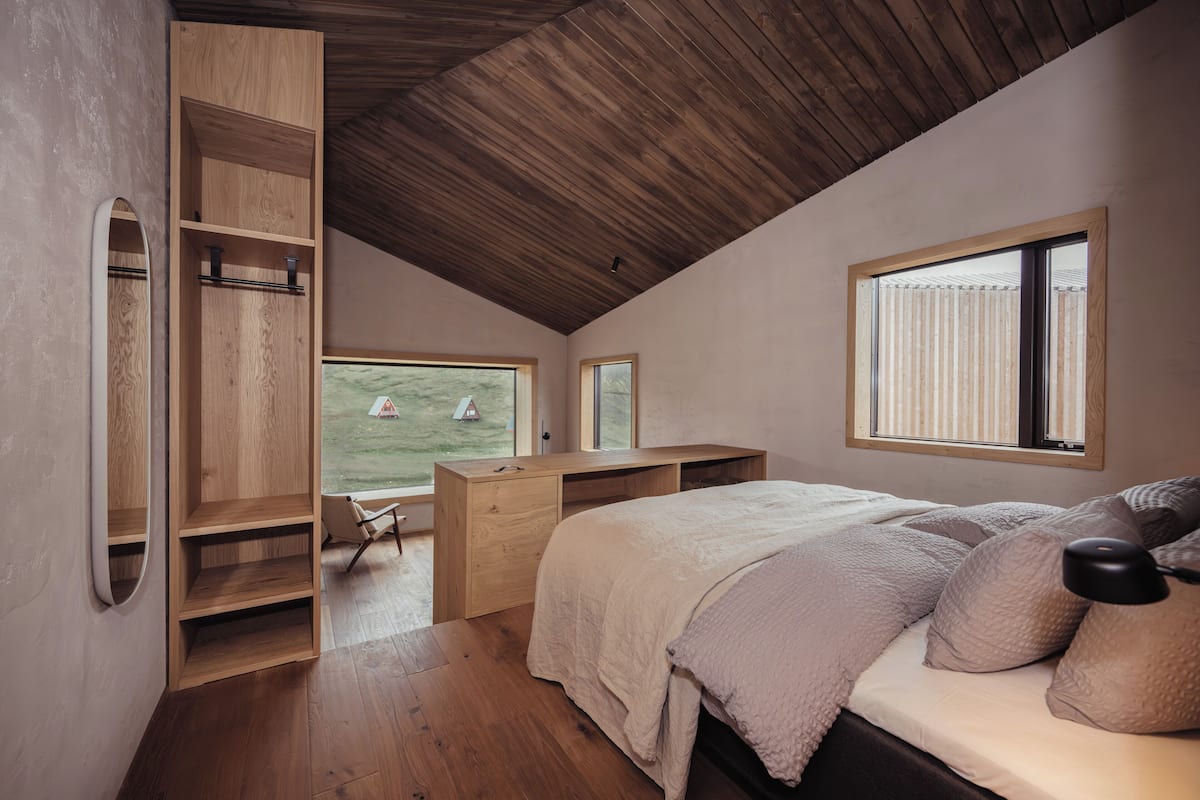 A bedroom with a sloped ceiling, wooden furniture, a bed with pillows, a desk, and a window overlooking a grassy area.