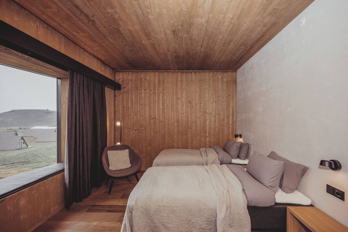 A bedroom with wooden walls, a large window, a bed, a chair, and a bedside table.