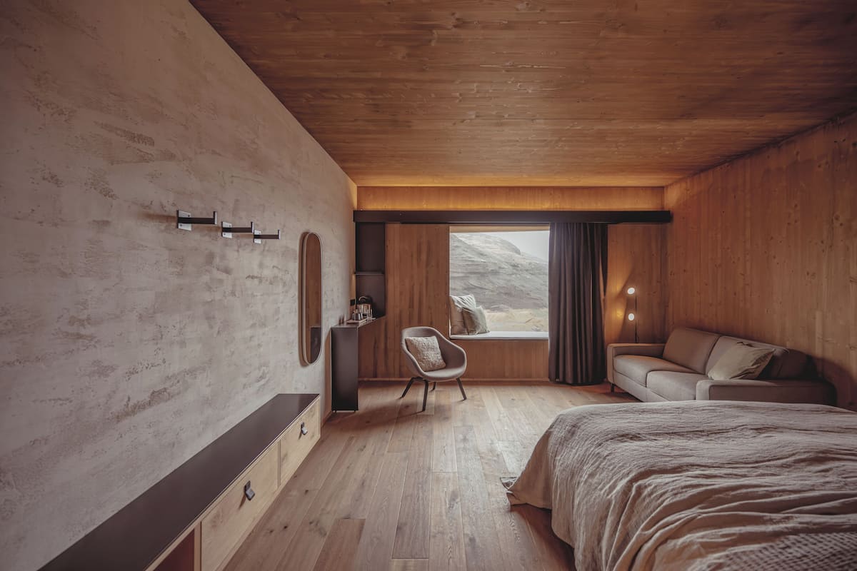 A bedroom with a wooden ceiling, a bed, a sofa, a chair, and a window with a view of a mountainous landscape.