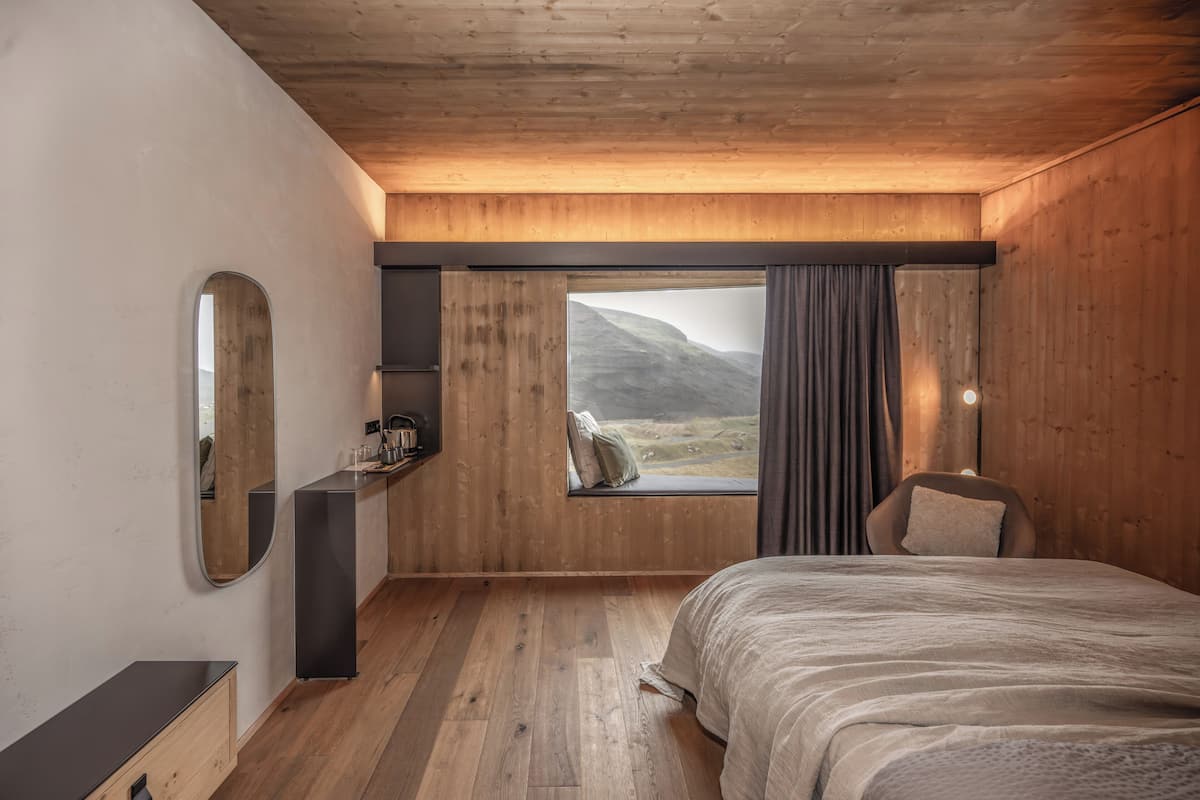 A bedroom with a wooden ceiling, a large window with curtains, a bed, a chair, and a mirror.