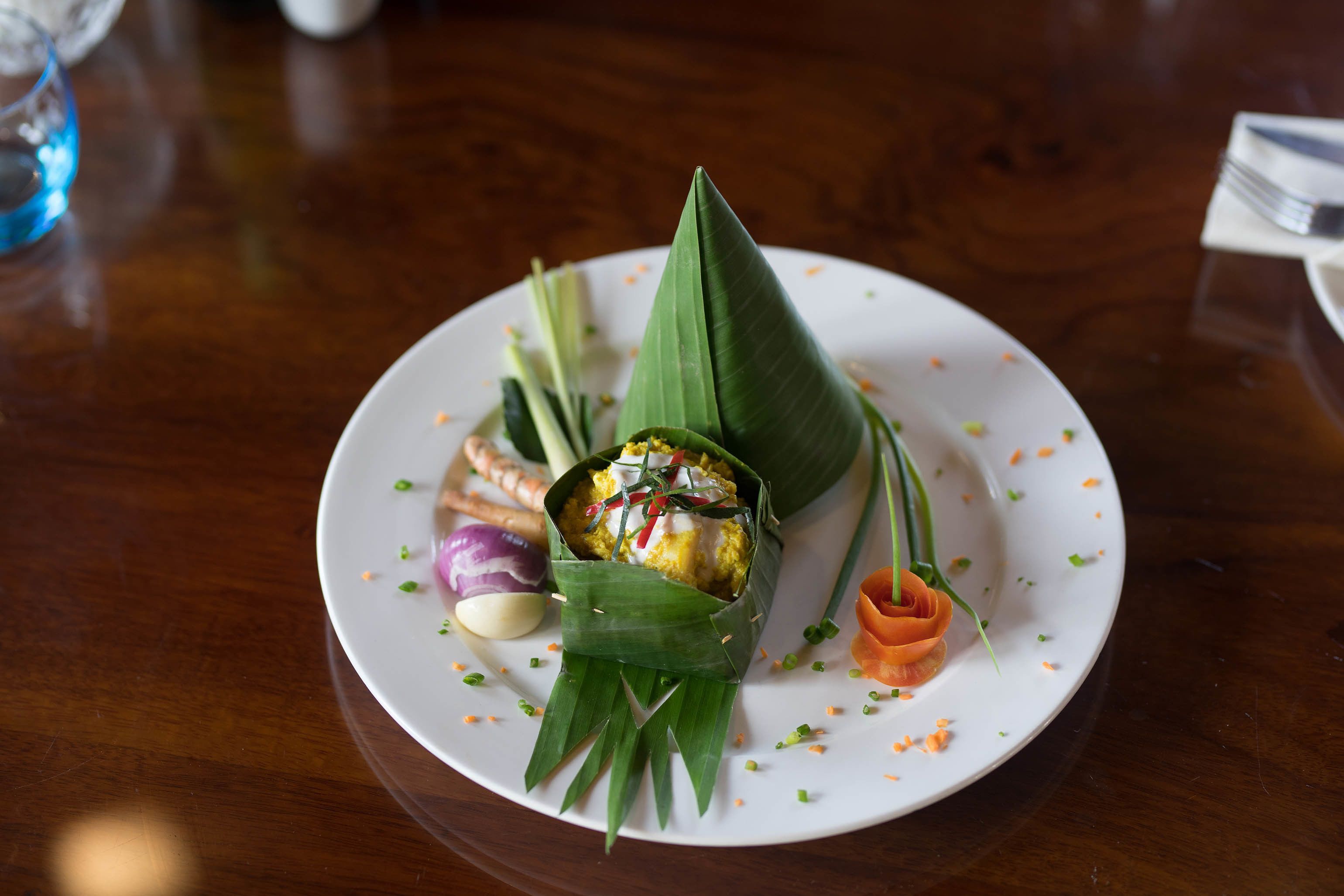 A plate with a food item wrapped in banana leaves, garnished with herbs and small vegetables.