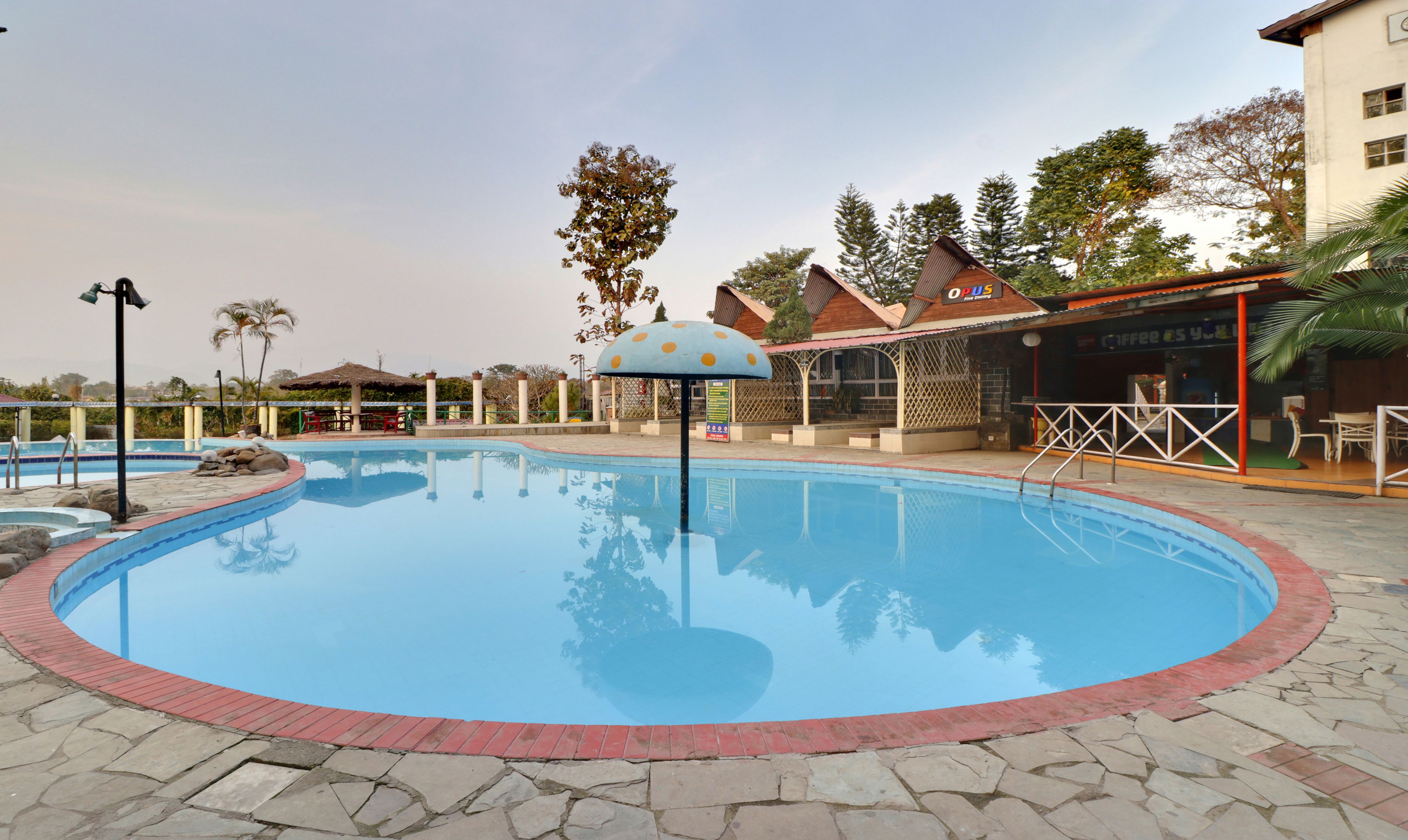 Une piscine entourée d’une terrasse en pierre, un espace salon couvert et un bâtiment portant une enseigne indiquant « OPUS, hôtel de première classe ».