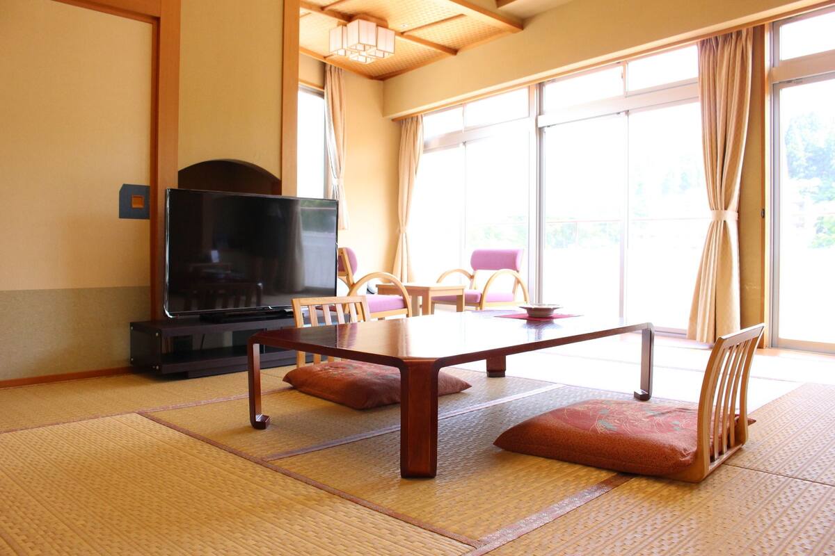 Una habitación japonesa tradicional con suelo de tatami, una mesa baja, sillas de madera, un televisor y ventanas grandes con cortinas.