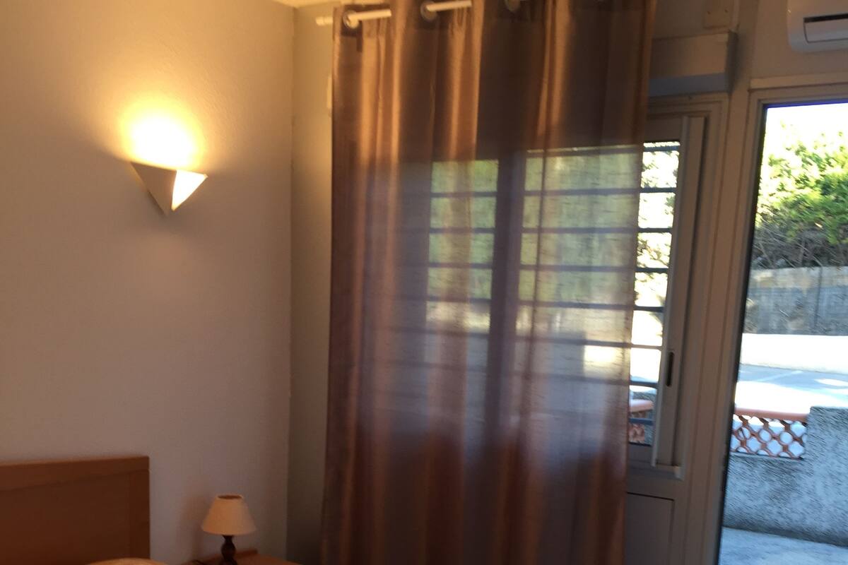A bedroom with a bed, a nightstand, a lamp, a window with curtains, and an air conditioning unit.