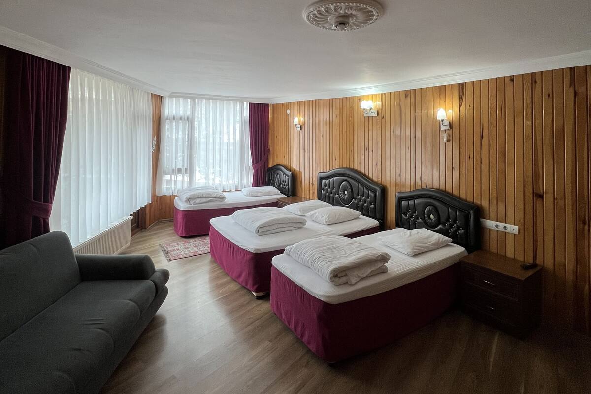 Quarto de hotel com duas camas, um sofá, painéis de madeira e uma janela grande com cortinas.