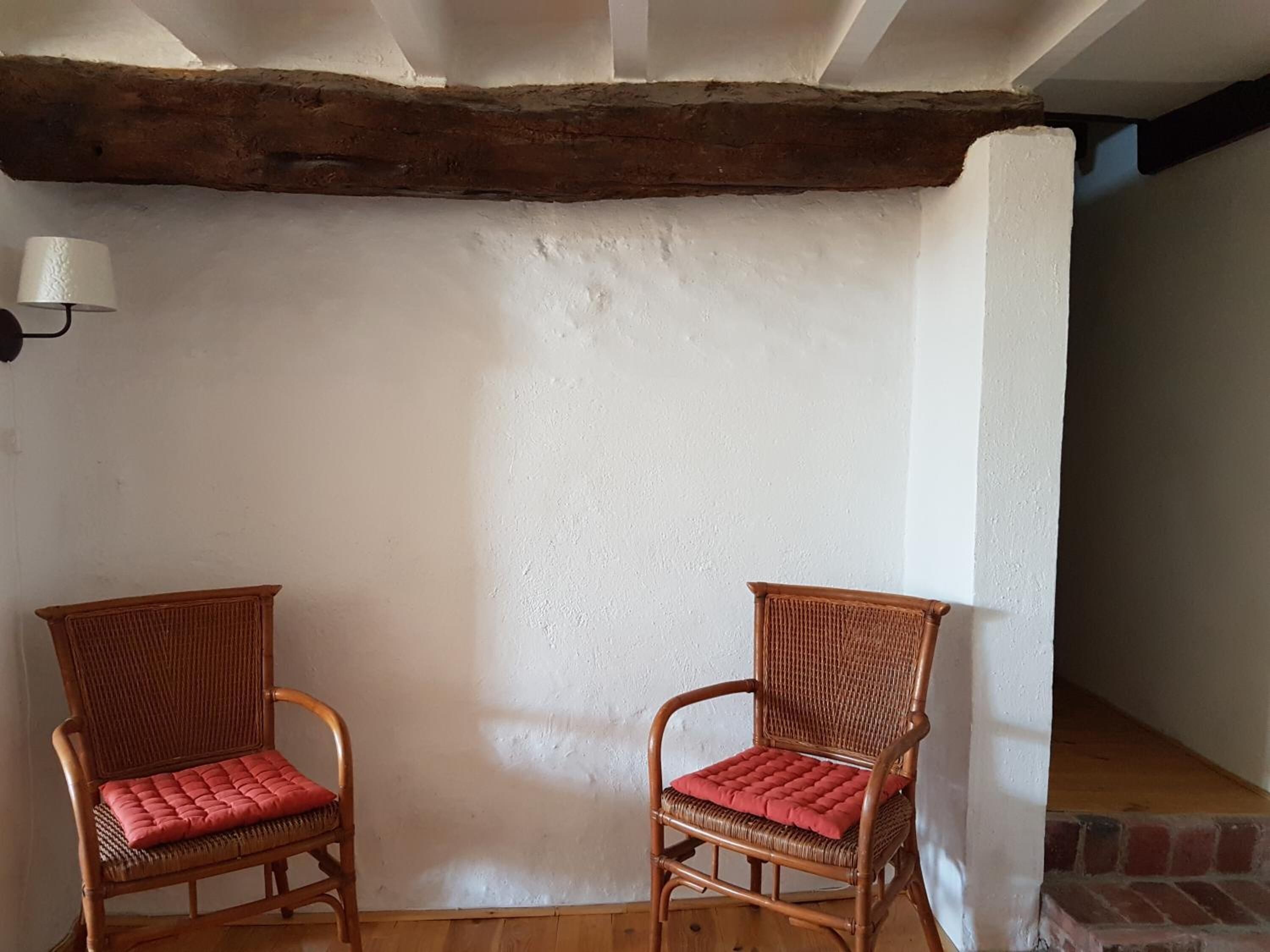 Deux fauteuils en osier avec des coussins rouges, dans une pièce aux murs blancs et aux poutres apparentes.