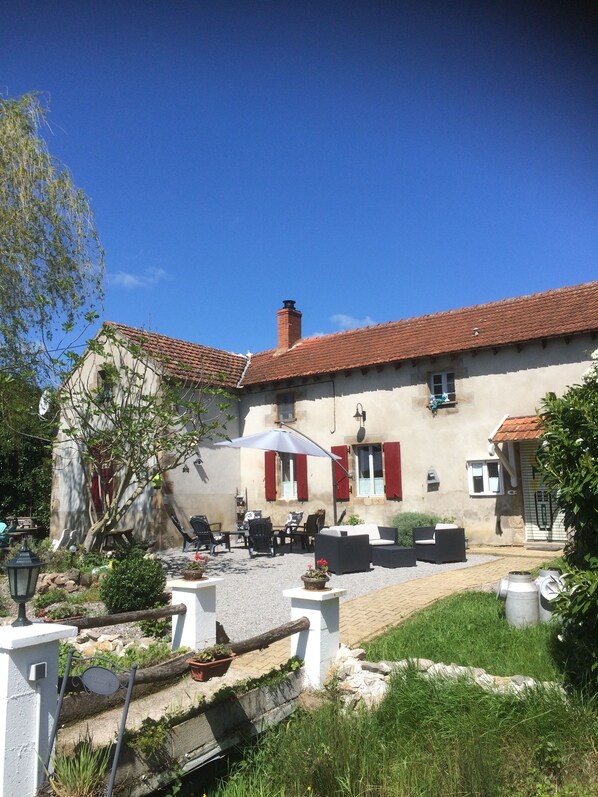A building with a red-tiled roof, outdoor seating area with chairs and tables, and a small garden with plants and a welcome sign.