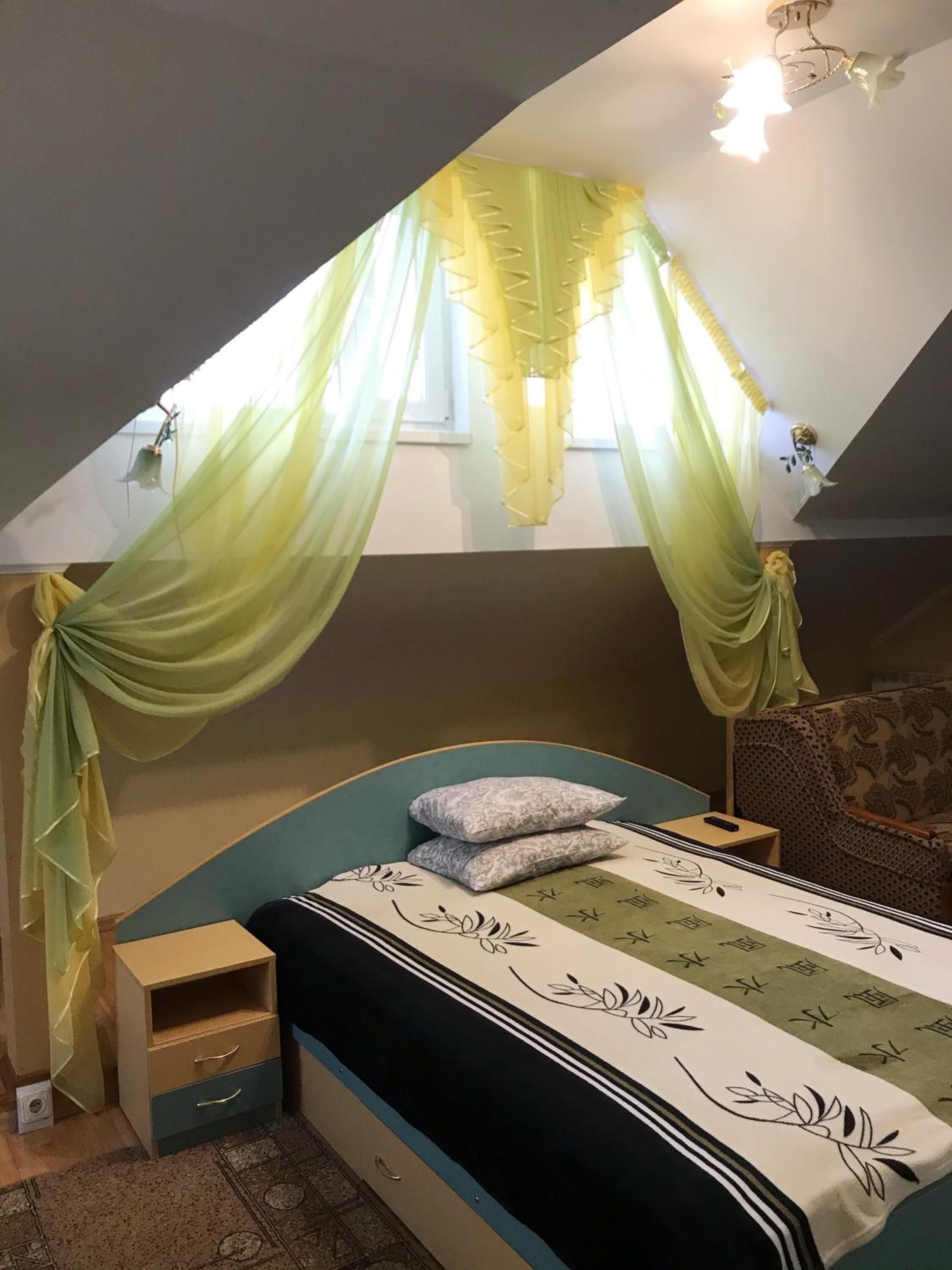 A bedroom with a bed, a nightstand, a chair, and a window with sheer curtains.