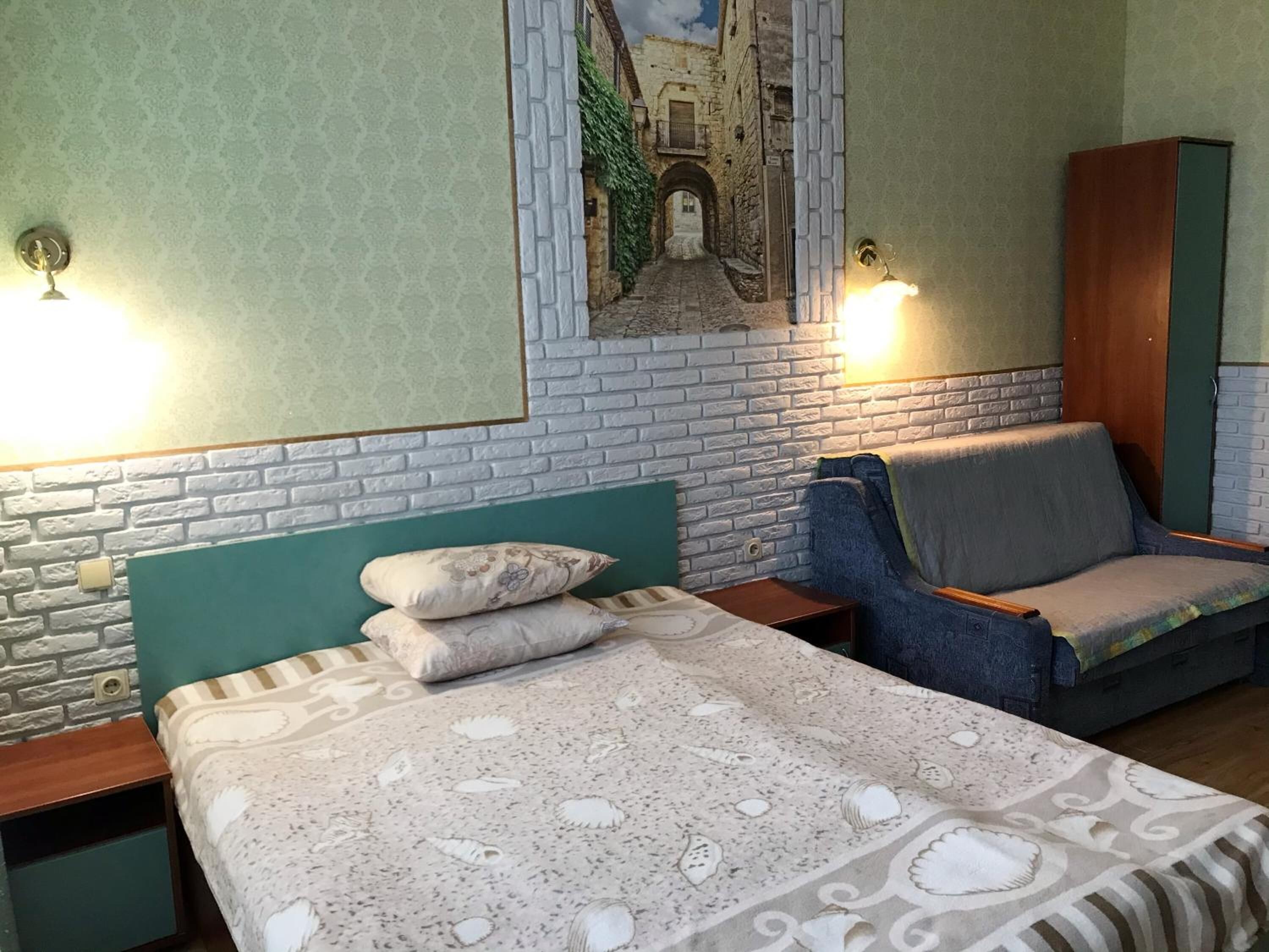 Una habitación con cama, una silla, una mesita de noche y una lámpara de pared.