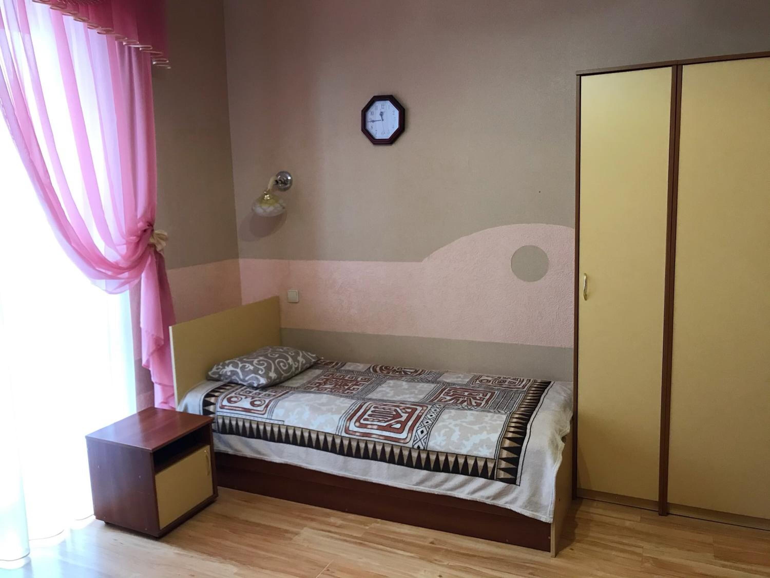 A bedroom with a bed, a nightstand, a wardrobe, a clock on the wall, and a window with pink curtains.