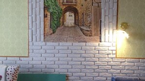 Une pièce décorée d’une fresque représentant une ruelle étroite, avec un lit recouvert d’une literie à motifs, une tête de lit verte, une petite table de chevet en bois et une chaise bleue.