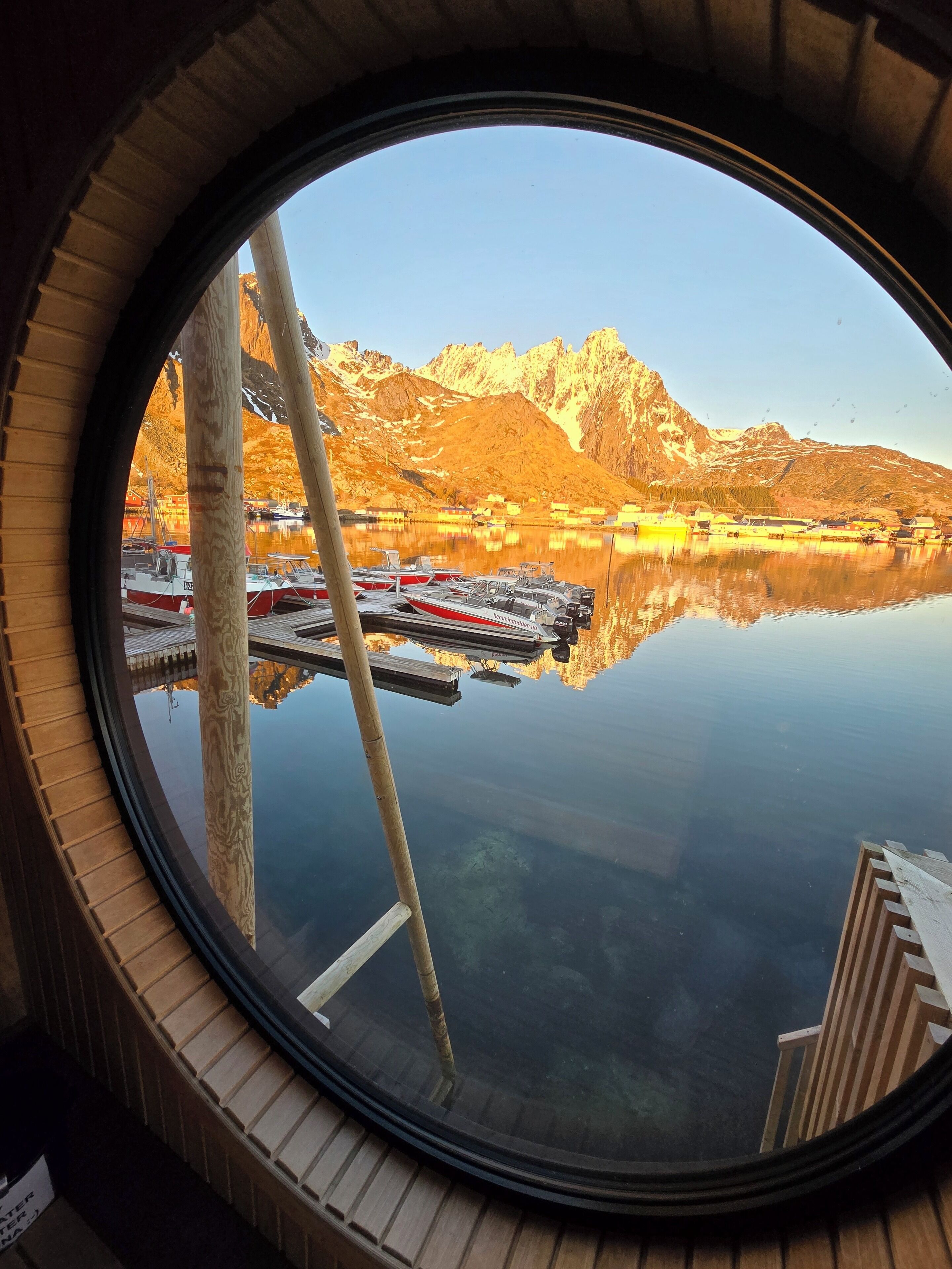 Ein rundes Fenster mit Blick auf einen stillen See, Boote und Berge.