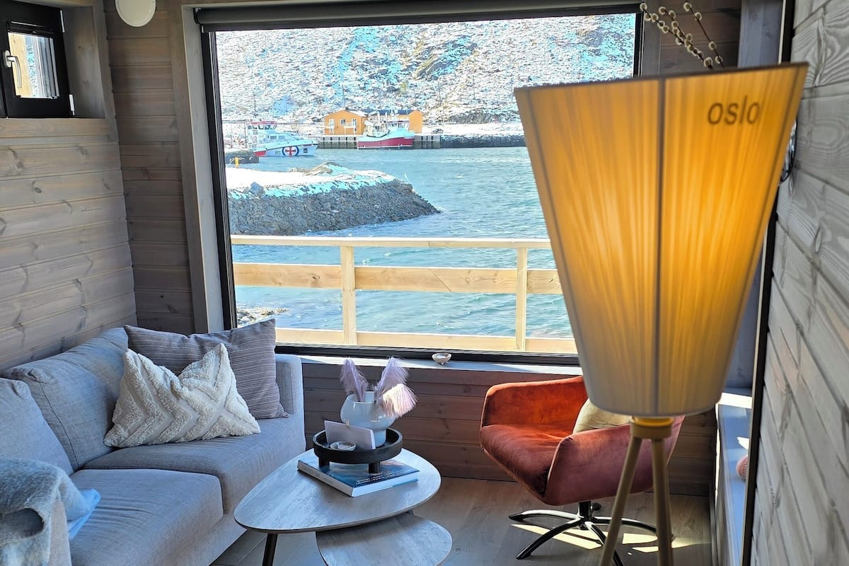 A modern living room with a sofa, a coffee table, and a red chair. A large window offers a view of a harbor with boats and buildings.