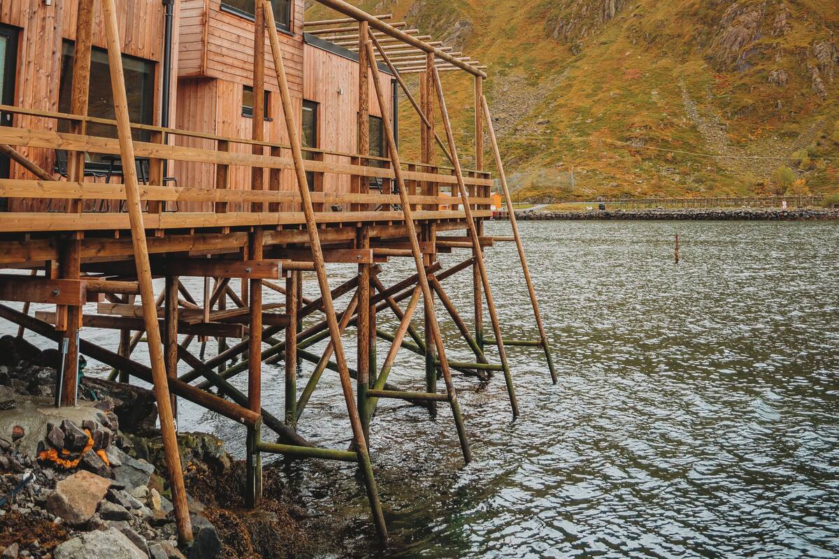 A wooden cabin on stilts by a lake with mountains in the background.