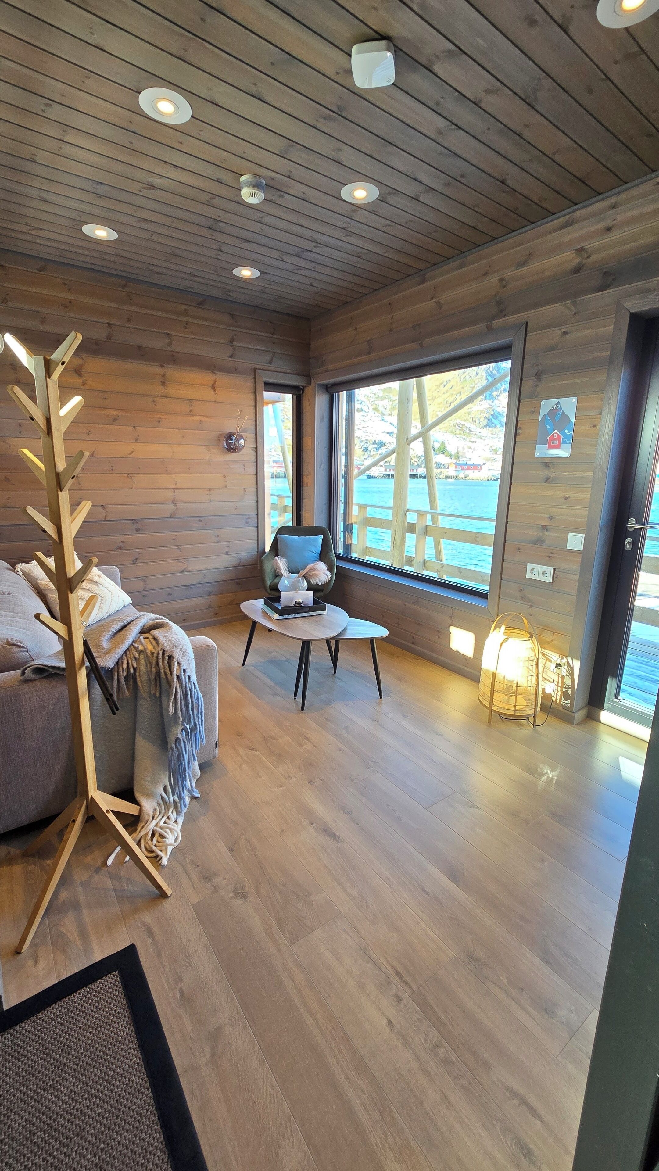 Una habitación acogedora con paredes y techo de madera, un ventanal con vistas al agua, un perchero de madera, un sillón cómodo y una mesita con un florero.