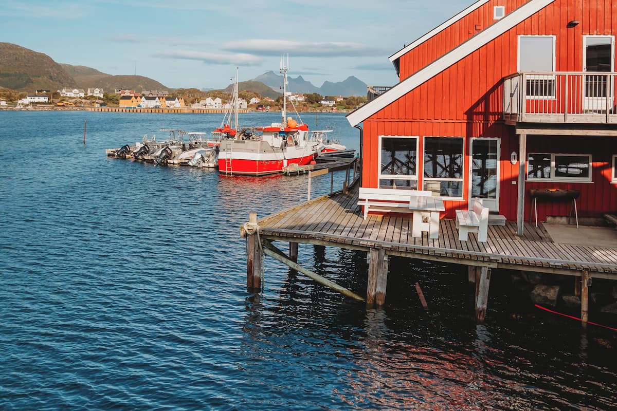 A red building with a dock extending into calm water, boats moored nearby, and a clear sky.