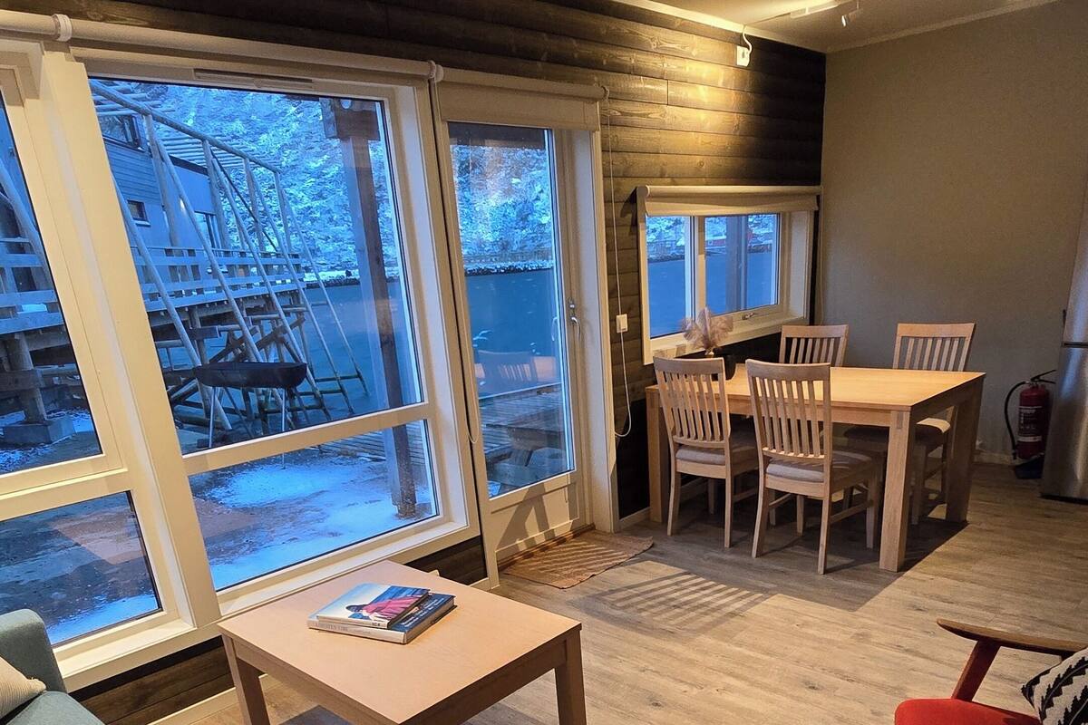 A modern living room with a sofa, a coffee table, and a dining area with chairs. Large windows provide a view of the outside.