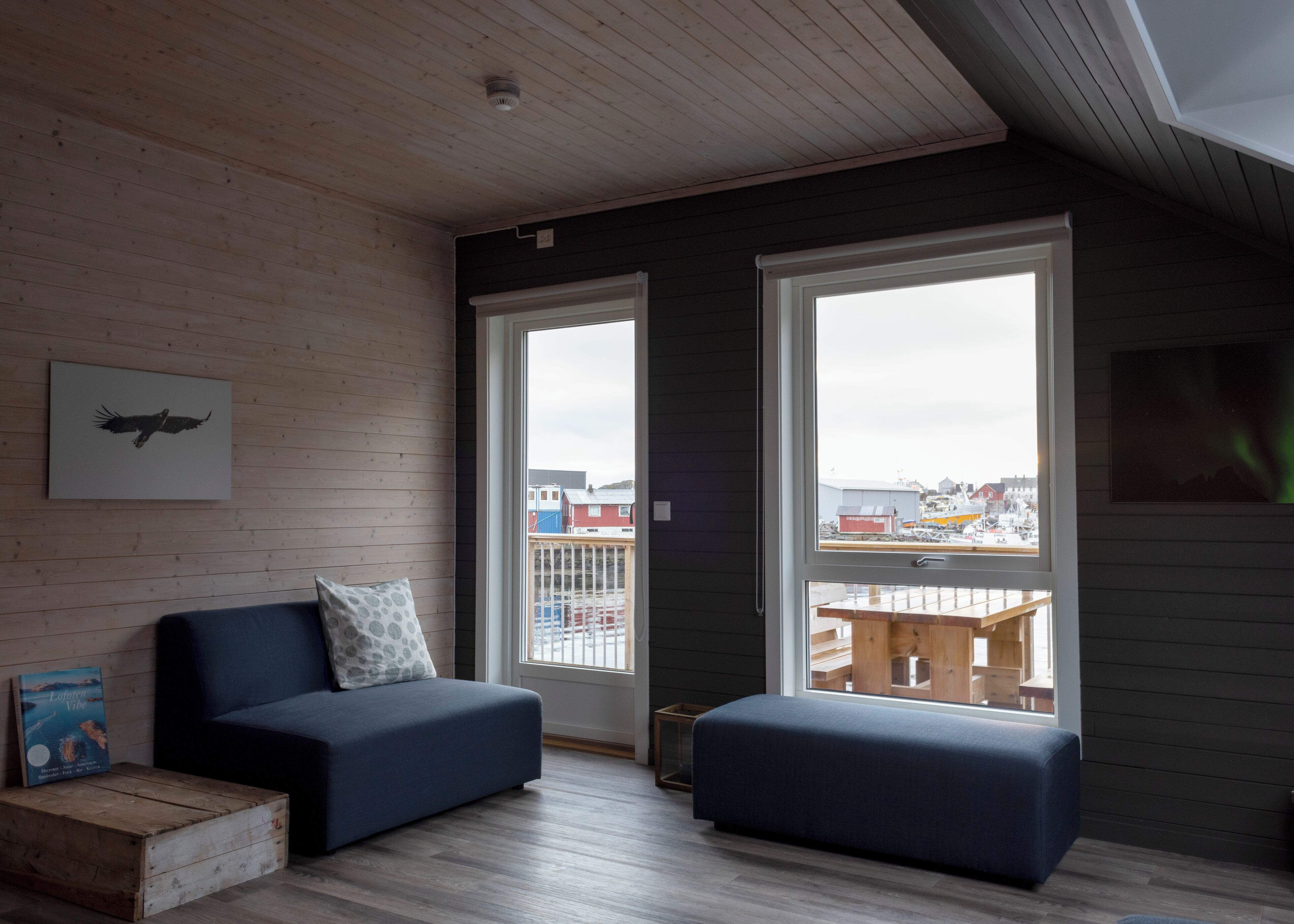 A modern living room with a dark blue sofa, a wooden bench, and a view of a dock and buildings through large windows.