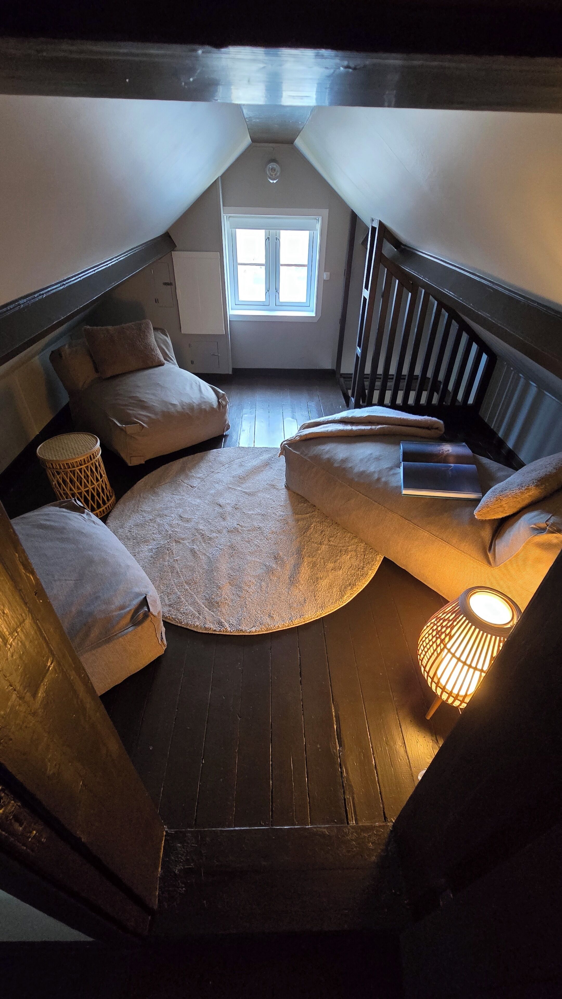 A loft bedroom with a sloped ceiling, a window, a circular rug, and a small lamp.