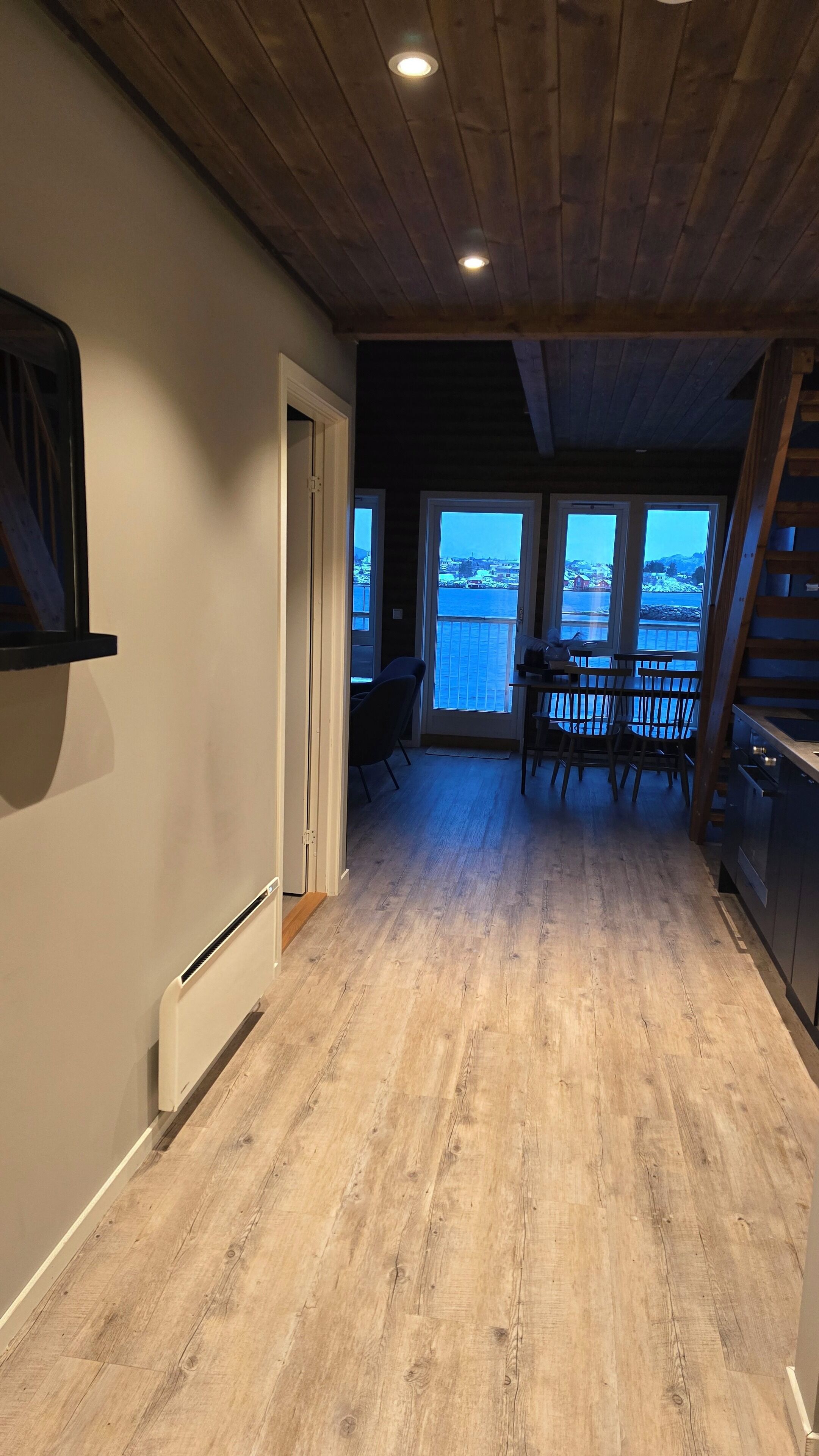 A spacious room with a wooden floor, a dining area with chairs and a table, and a view through large windows.