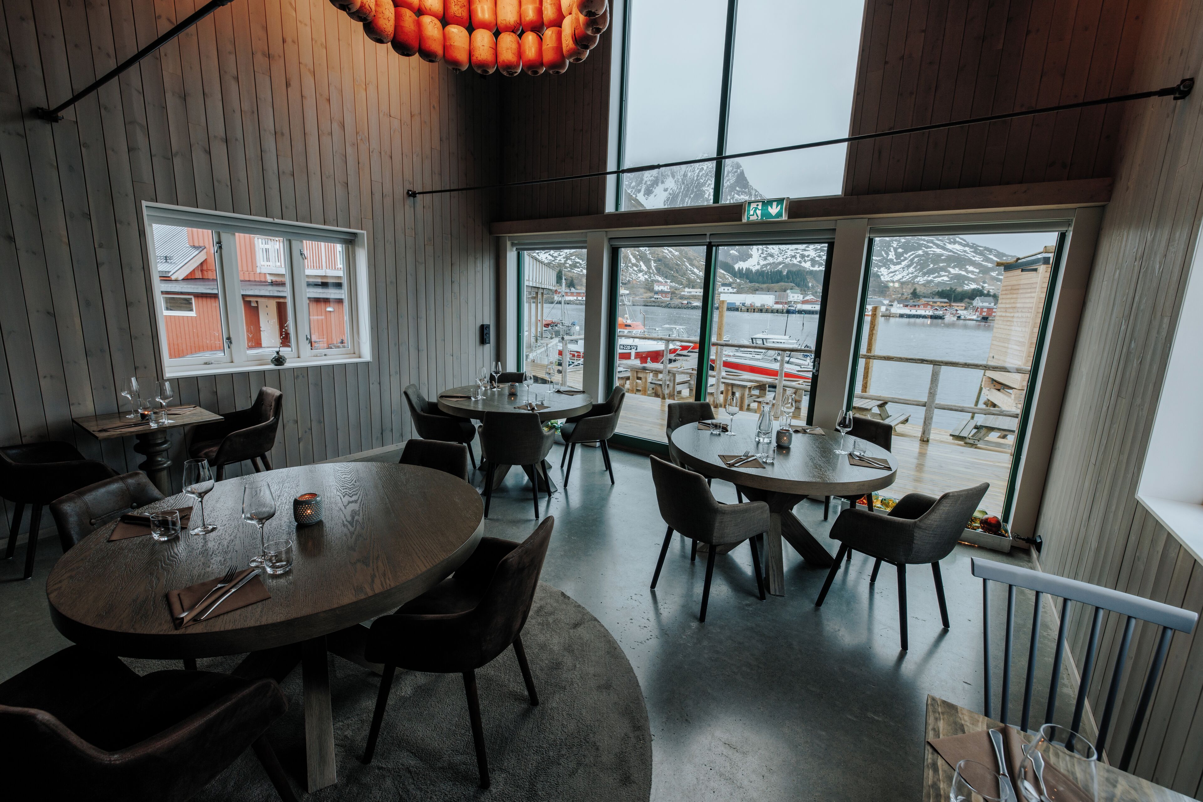 Ein Restaurant mit runden Tischen und Stühlen, großen Fenstern mit Blick auf einen Hafen und Berge, sowie Holzwänden.