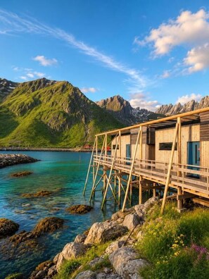 A wooden structure with a deck extending over water, surrounded by mountains and clear skies. - Hemmingodden Lodge (Vestvågøy)
