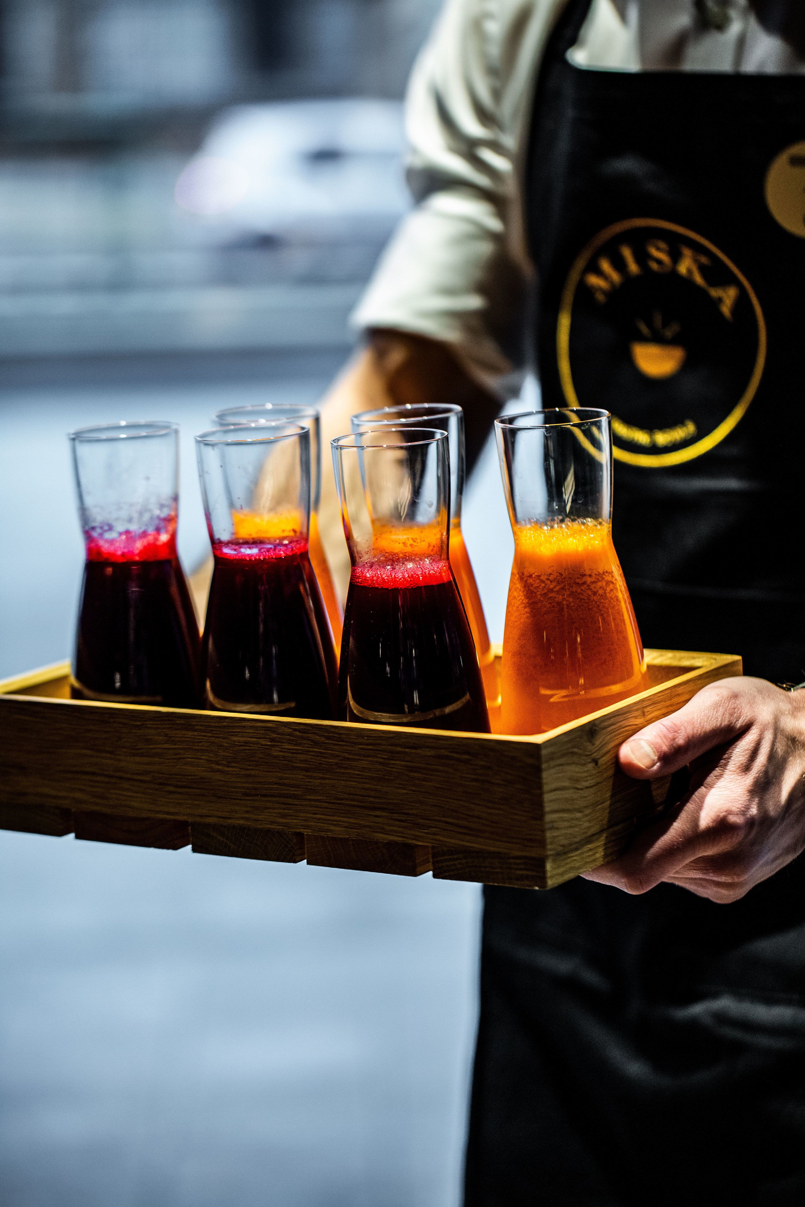 A person holding a wooden tray with four glasses of different colored beverages.