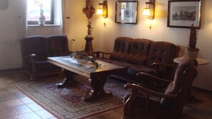 Un salon comprenant un canapé, deux fauteuils, une table basse et un tapis. Des lampes murales et des tableaux encadrés ornent les murs.