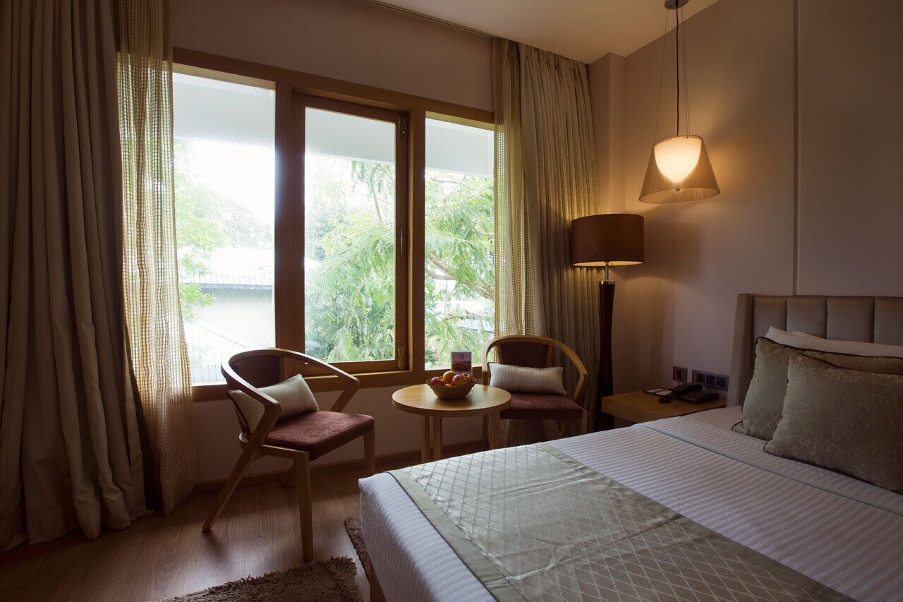 Habitación de hotel con cama, dos sillas, una mesita con frutero, una lámpara y vistas a zonas verdes por la ventana.