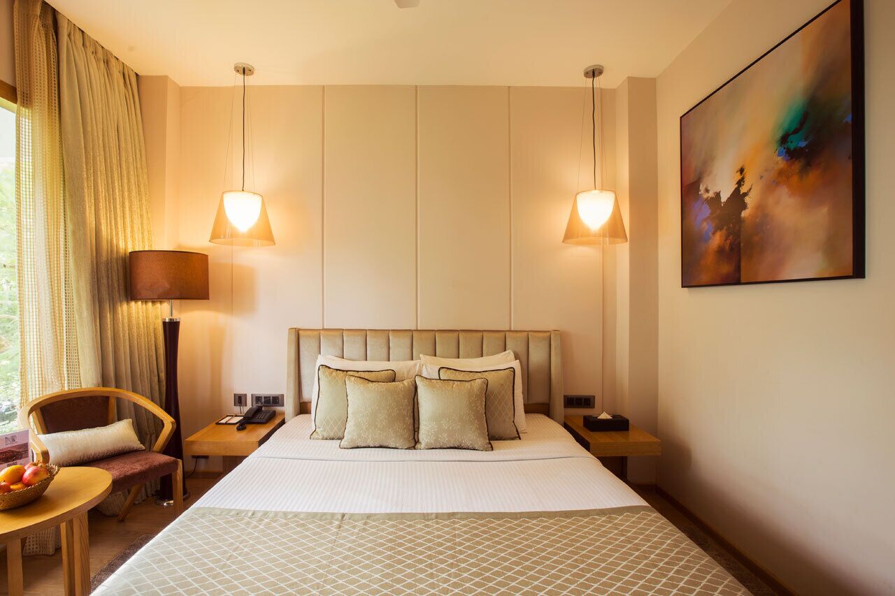A hotel room with a large bed, two bedside lamps, a nightstand, a chair, a table with fruit, and a painting on the wall.