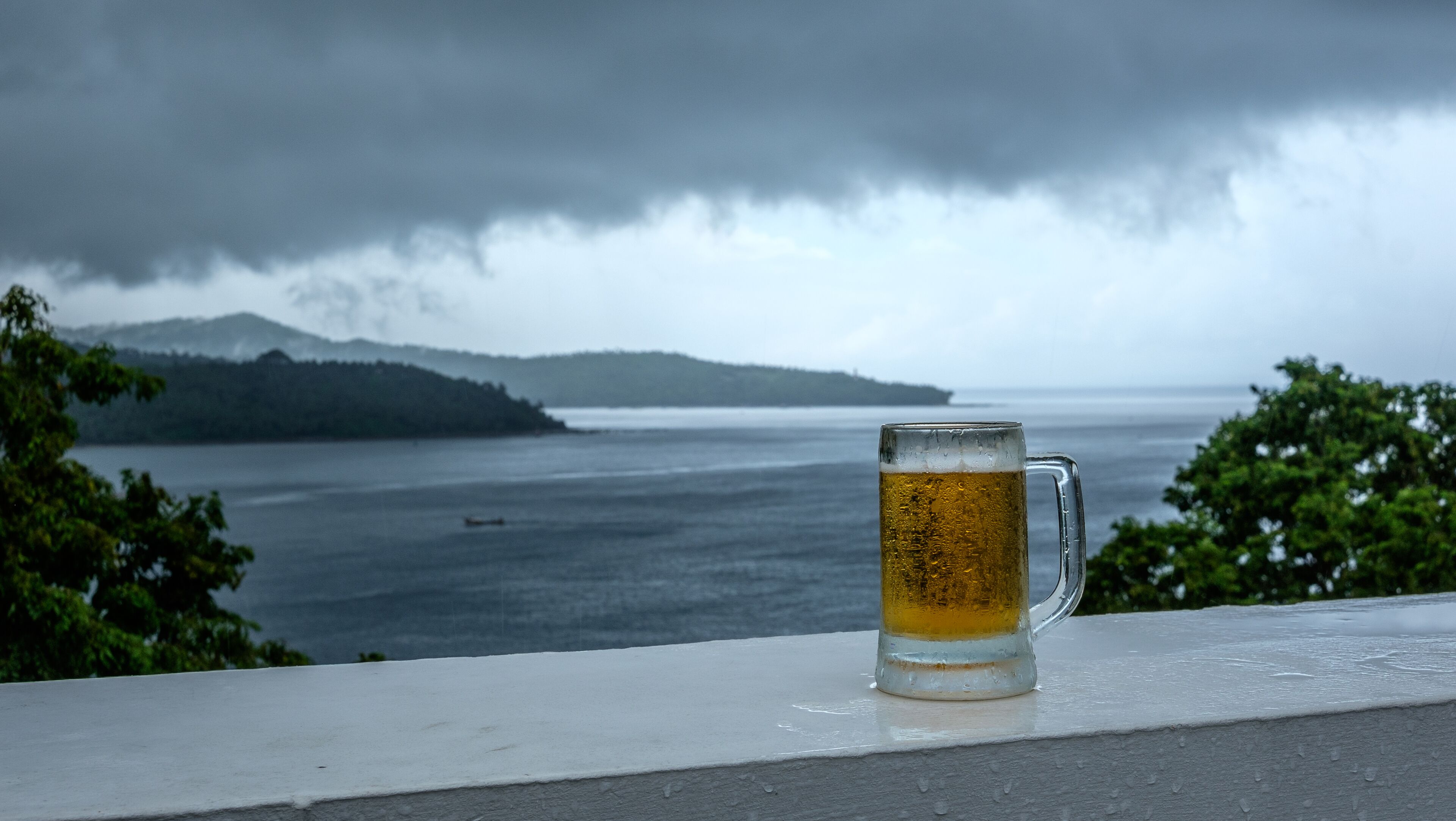 A glass of beer on a ledge with a view of a body of water and mountains in the background.