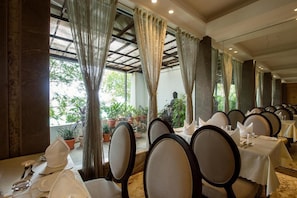 Un restaurante con varias mesas dispuestas para comer, amplias ventanas con cortinas y plantas en macetas.