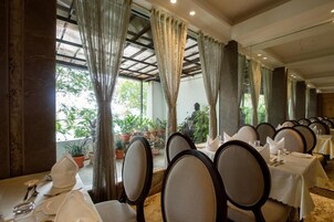Un restaurante con varias mesas dispuestas para comer, amplias ventanas con cortinas y plantas en macetas.
