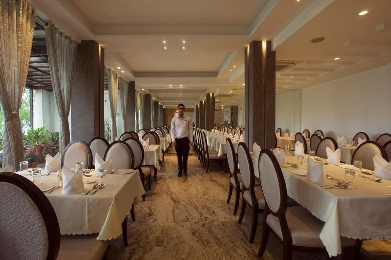 A long corridor with tables set for dining, a waiter walking down the center, and large windows on one side.
