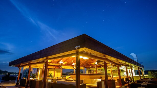 Un área de estar al aire libre techada, con sillas y mesas, bajo una estructura de techo de madera, con vista al cielo nocturno.