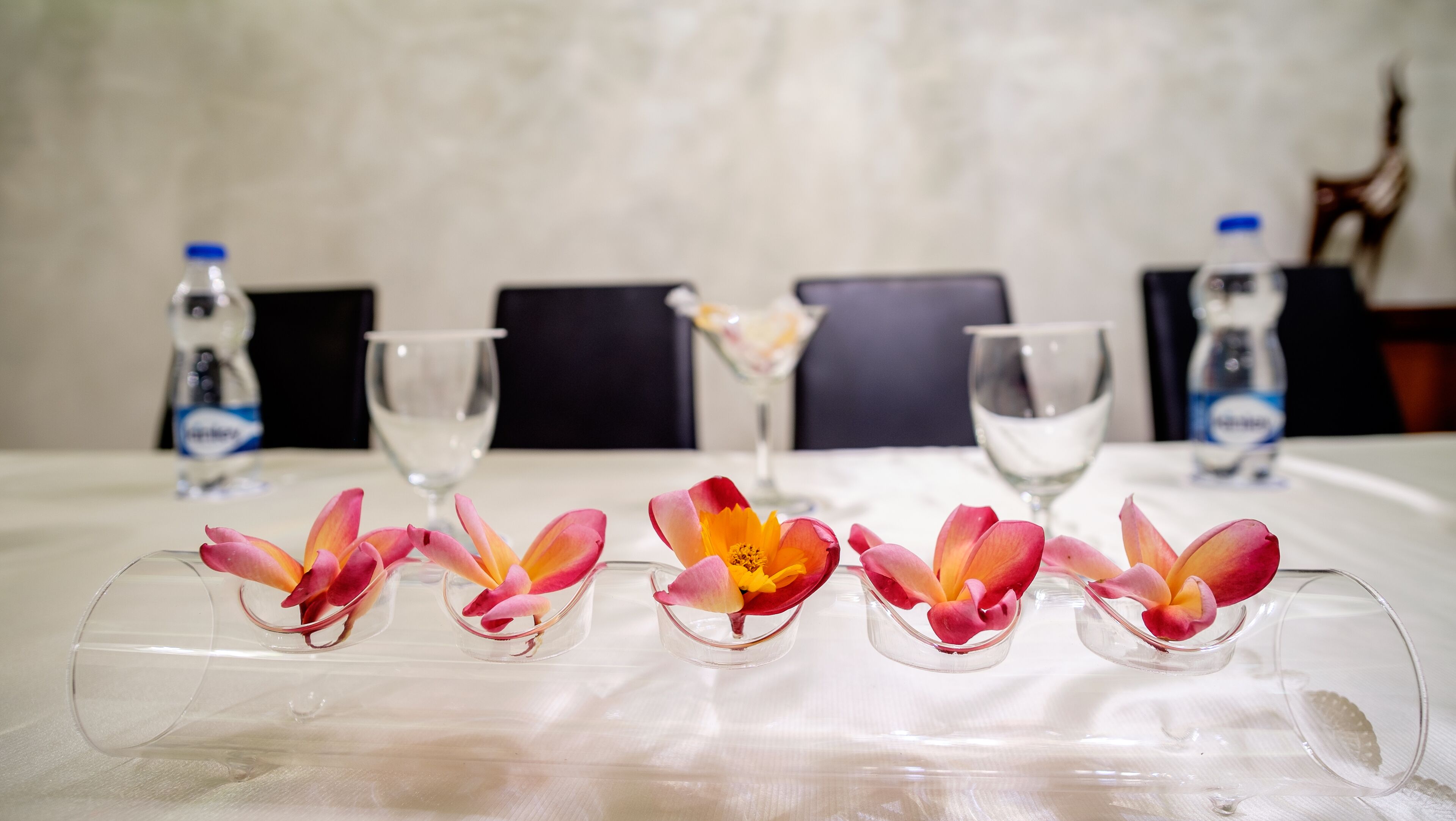 Een tafel met transparante glazen vazen gevuld met roze en gele bloemen, waterflessen en lege wijnglazen.