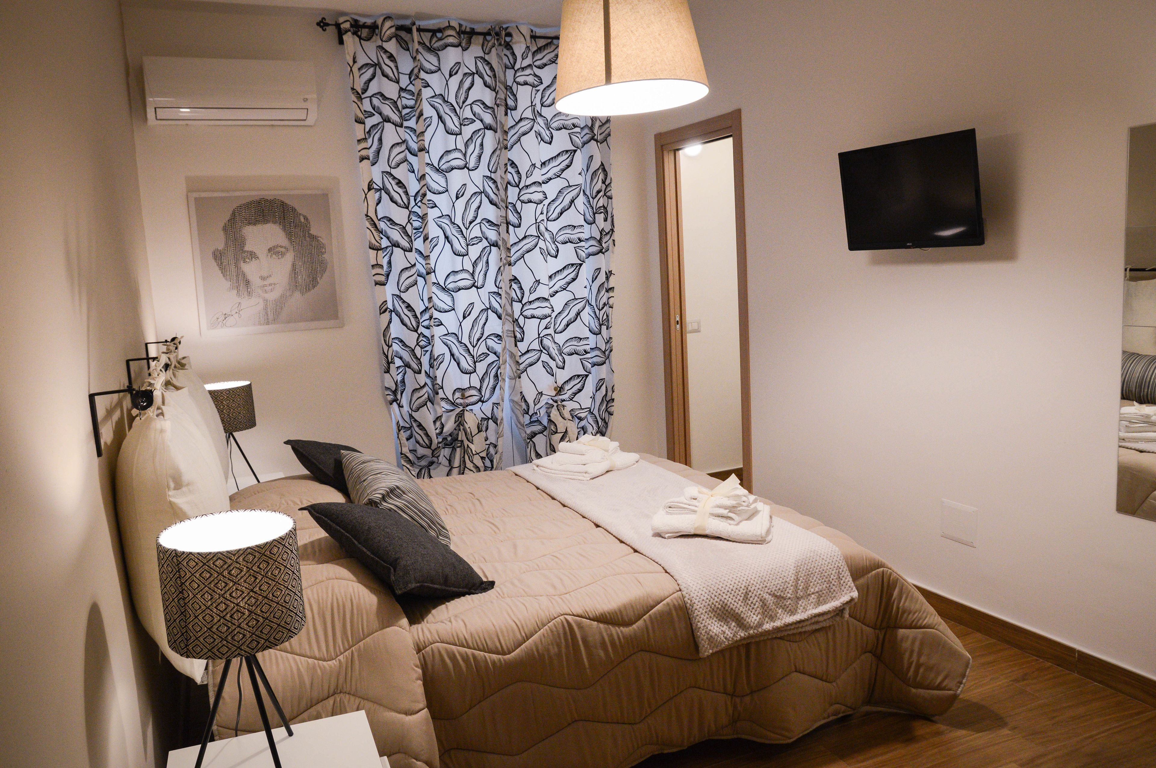 A neatly made bed with a beige duvet, a patterned throw blanket, and a folded towel. There is a bedside lamp with a geometric pattern, a wall-mounted TV, and a framed picture on the wall.