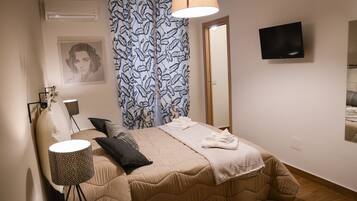 A neatly made bed with a beige duvet, a patterned throw blanket, and a folded towel. There is a bedside lamp with a geometric pattern, a wall-mounted TV, and a framed picture on the wall.