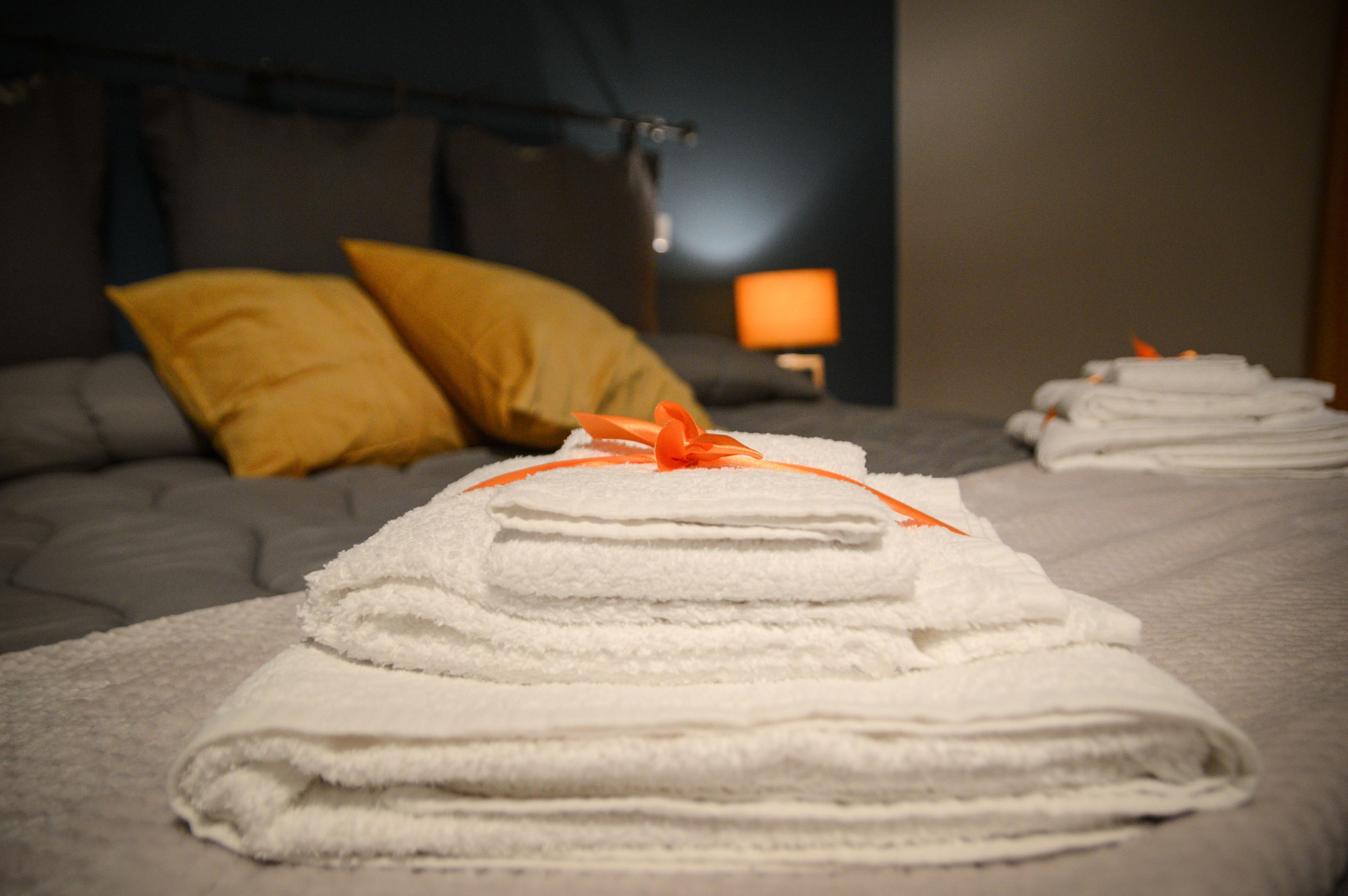 A neatly made bed with a stack of folded white towels tied with an orange ribbon, a yellow pillow, and a bedside lamp.