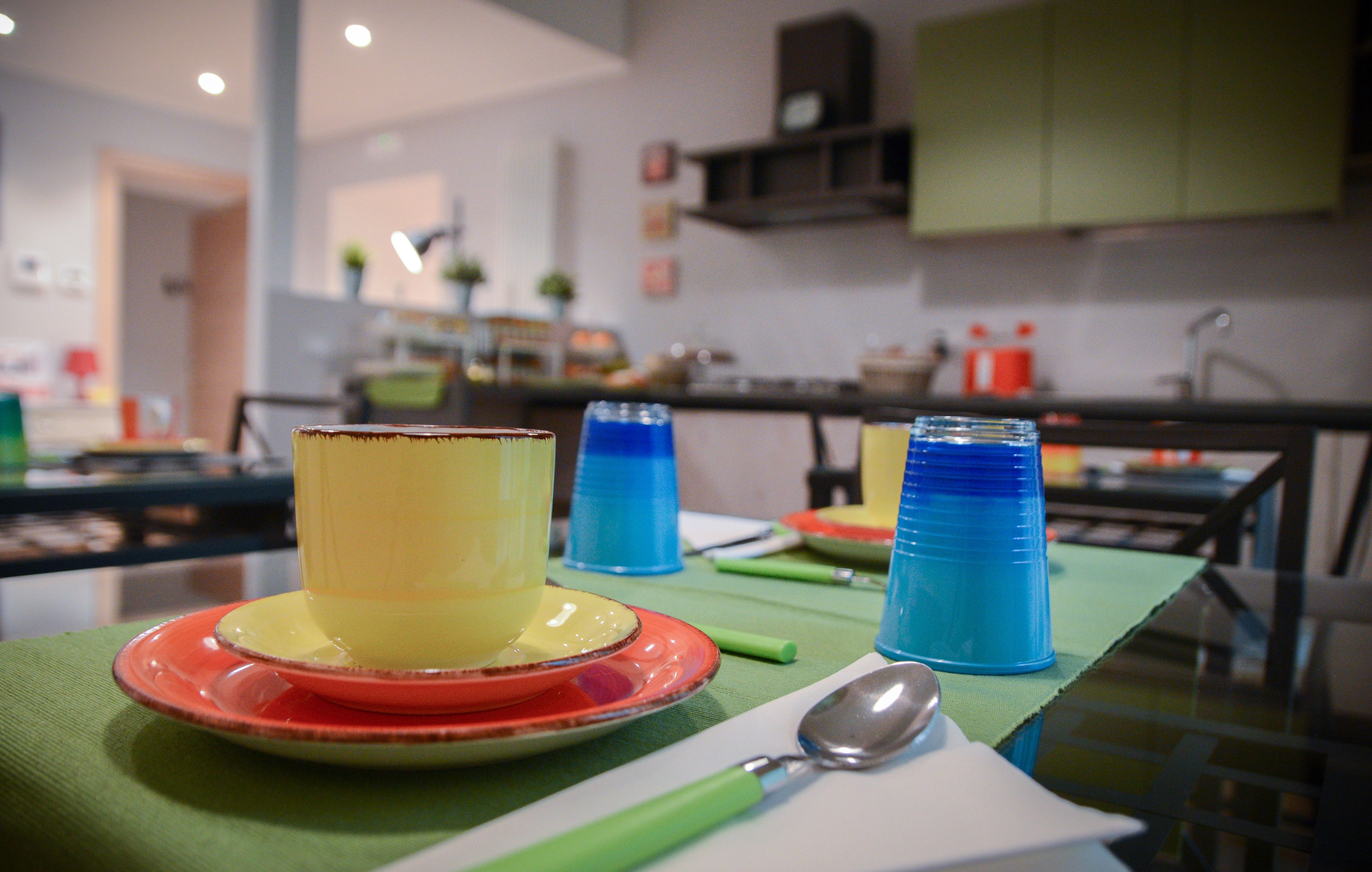 A dining table with a yellow cup on a red plate, a blue cup, and a spoon.