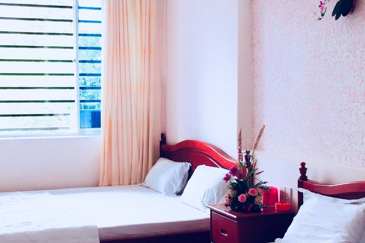 Habitación de hotel con dos camas, una ventana con cortinas, un arreglo floral en la pared y una mesita de noche con un florero.