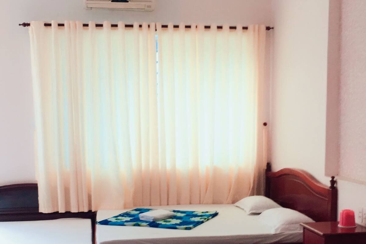 Habitación de hotel con dos camas, cabecero de madera, mesita de noche y ventana con cortinas traslúcidas.
