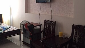 A living room with a television mounted on the wall, a dining table with chairs, a standing fan, and a bed with a patterned bedspread. - Quang Diep Hostel (Ho Chi Minh City)